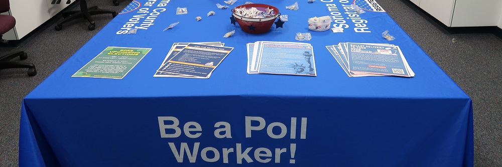 A picture looking down at a large table with a blue tablecloth over it that says "Be a Poll Worker!" in large letters on the side. On top of the table there are four piles of different kinds of fliers, a bowl of mints, "I Voted" stickers, and old election pins. 