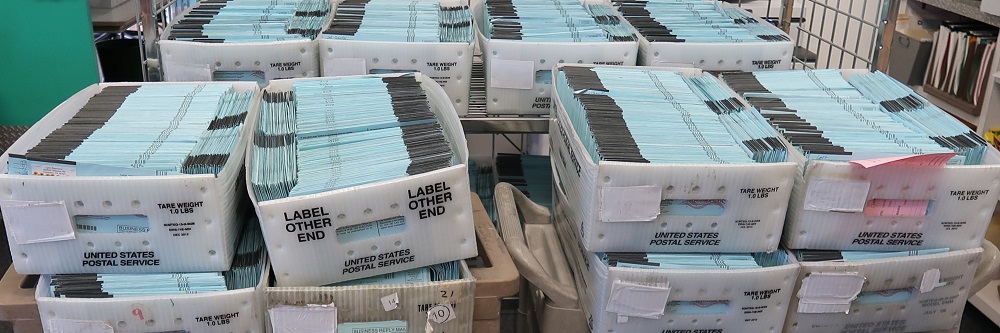 A picture of multiple mail trays containing hundreds of blue Vote-by-Mail ballot envelopes